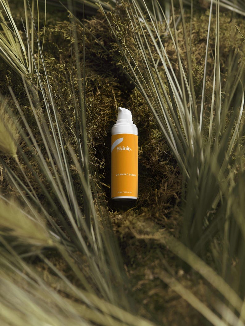 K&ouml;nnte beinhalten: Eine Hautpflegeproduktflasche mit wei&szlig;em Pumpkopf und orangefarbenem Etikett. Auf dem Etikett steht "Skinity" und "Vitamin C Serum". Die Flasche befindet sich vor einem Hintergrund aus gr&uuml;nem Laub.