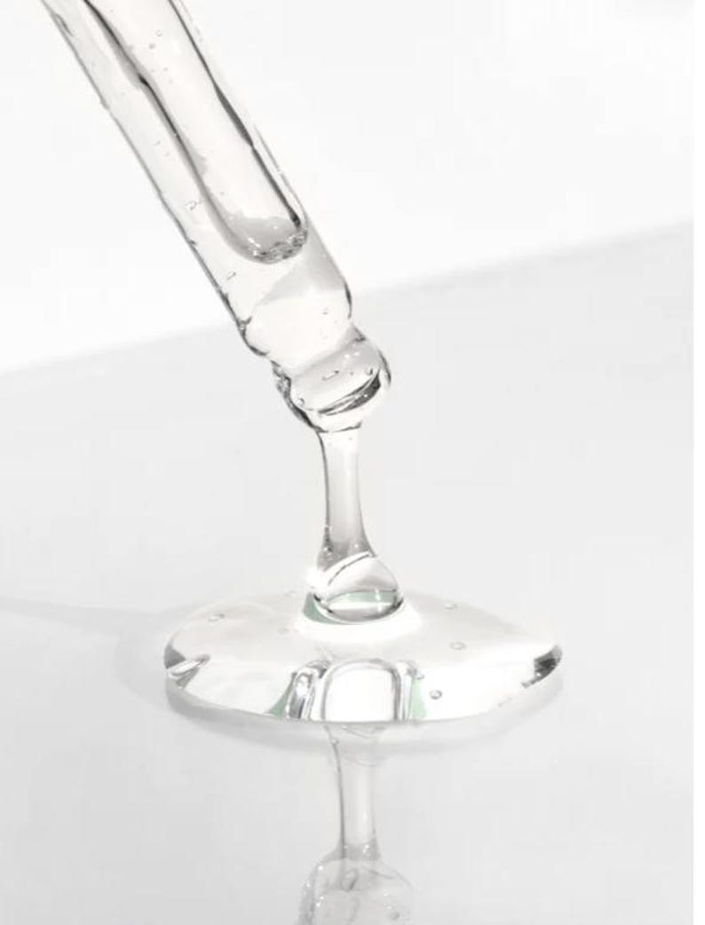 K&ouml;nnte beinhalten: Nahaufnahme einer transparenten Glaspipette, die eine z&auml;hfl&uuml;ssige, transparente Fl&uuml;ssigkeit abgibt. Die Fl&uuml;ssigkeit bildet eine kleine, flache Pf&uuml;tze mit reflektierender Oberfl&auml;che. Der Hintergrund ist hell und wei&szlig;, was die Klarheit des Produkts hervorhebt.