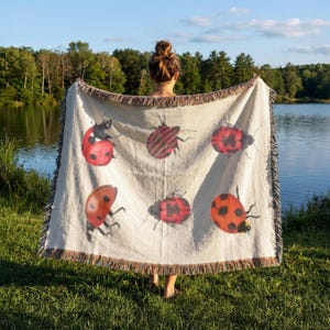 May include: A cream-colored woven throw blanket featuring a repeating pattern of six red ladybugs with black spots. The blanket has a textured fabric construction with frayed edges along the top and bottom, displaying a variety of beetle illustrations.