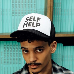 May include: A white trucker hat with the words "SELF HELP" in bold black letters. The hat has a black brim and a black mesh back. The hat is worn by a person wearing a black and white plaid shirt.