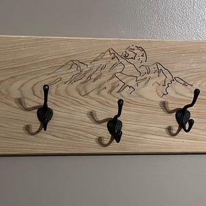 Puede incluir: Perchero de pared de madera clara con cinco ganchos de metal negro. La madera presenta un diseño de cordillera grabado. Los ganchos están espaciados uniformemente a lo largo del borde inferior.