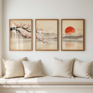 May include: A set of three framed wall art prints featuring a landscape with a calm lake, distant mountains, and cherry blossom branches with pink flowers. The third print displays a large red sun above the horizon, all in wooden frames.
