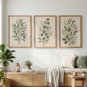 Op de afbeelding: Een set van drie ingelijste wandkunstprints met botanische illustraties van groene bladeren en olijftakken op een lichtbeige achtergrond, elk in een eenvoudige lichthouten lijst en horizontaal gerangschikt aan een witte muur.
