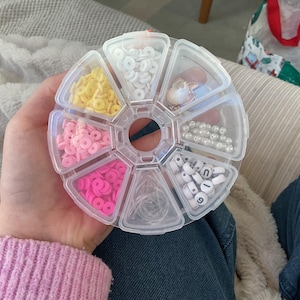 Könnte beinhalten: Ein klarer, segmentierter Behälter gefüllt mit verschiedenen Bastelmaterialien. Der Behälter enthält gelbe, weiße, rosa und pinkfarbene Polymer Clay Perlen, weiße Buchstabenperlen, Perlen und transparentes elastisches Band.