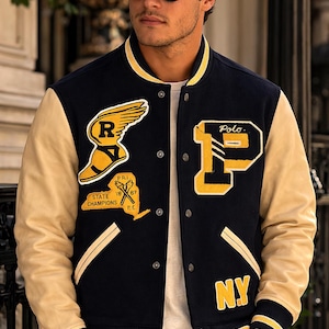 May include: Navy blue and tan varsity jacket with yellow accents. Features a large "P" and "NY" patch, along with other patches. The jacket has a ribbed collar, cuffs, and hem. The sleeves are tan, and the body is navy blue.