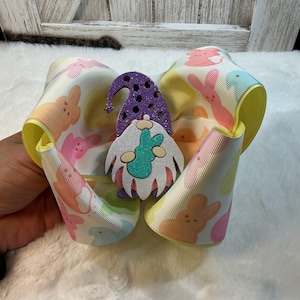 May include: A large, pastel-coloured hair bow with a repeating pattern of Easter bunnies. The bow has yellow edges and a glittery purple gnome embellishment holding a turquoise egg. The bow is on a white, fluffy surface.