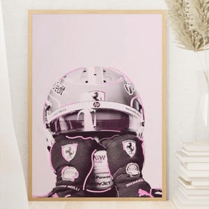 Puede incluir: Impresión enmarcada que presenta un casco y guantes de piloto de Fórmula 1, sobre un fondo rosa. El casco muestra logotipos de patrocinadores y los guantes tienen el logotipo de Ferrari. La obra está en un marco de madera.