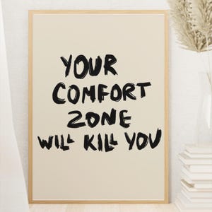 May include: A framed print with the text "YOUR COMFORT ZONE WILL KILL YOU" in bold black lettering on a cream background. The frame is a light wood color. The print is displayed near a vase with dried plants and a stack of books.