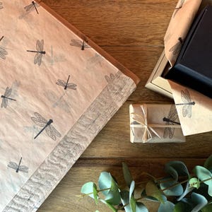 May include: Overhead view of gift packaging featuring a dragonfly print on brown paper. A black box is open, revealing a dark interior. A small wrapped package is tied with twine. The items are on a wooden surface.