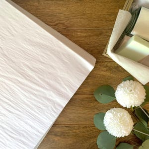 May include: A large sheet of white crinkled tissue paper positioned next to a wooden box containing two rectangular bars of soap wrapped in white paper, accompanied by two white pom-pom flowers with green eucalyptus leaves.