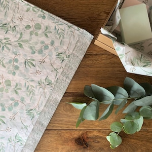 May include: A sheet of white tissue paper featuring a repeating pattern of green eucalyptus leaves and branches, partially covering a cardboard box containing a light green rectangular object, with a sprig of fresh eucalyptus leaves nearby.
