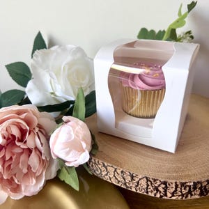 May include: A single cupcake with mauve frosting and small sprinkles inside a white cardboard box with a clear plastic window. The cupcake sits in a gold metallic liner. The box is positioned next to artificial white and light pink roses.