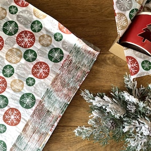 May include: A stack of white tissue paper featuring a repeating pattern of red, green, and gold circles with white snowflake designs. A red mug with a tree cutout sits in a cardboard box, accompanied by a frosted evergreen branch.