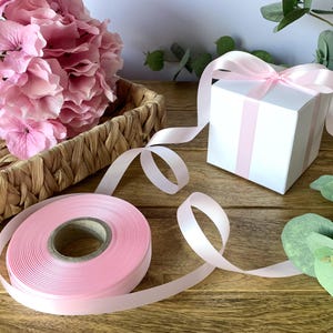 May include: A spool of pink satin ribbon is next to a white gift box with pink ribbon detailing and a pink bow. A woven basket of pink hydrangeas is in the background. The items are on a wooden surface.