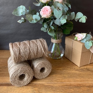 May include: Three cylindrical spools of natural brown jute twine arranged in a cluster. A clear glass vase holds a bouquet of light pink roses and eucalyptus leaves, and a square cardboard gift box is tied with a matching piece of twine.