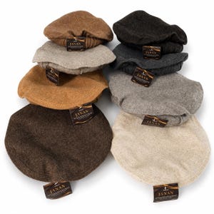 Op de afbeelding: Een collectie van acht platte petten in diverse neutrale tinten, waaronder bruin, antraciet, grijs, beige en tan. Elke pet heeft een textuur die op wol lijkt en een klein rechthoekig label aan de klep met de merknaam JANAN erop gedrukt.