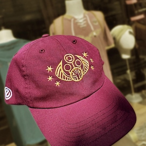 Puede incluir: Gorra de béisbol burdeos con visera curva. La gorra presenta un diseño dorado bordado de una cara estilizada con acentos de estrellas. Un logotipo blanco está en el lateral.