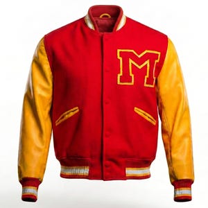 Op de afbeelding: Een rode varsity jas met gele mouwen en een wit en geel gestreepte zoom en manchetten. De jas heeft een grote gele letter "M" op de borst en twee voorzakken.