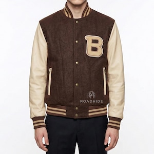 May include: A brown and cream varsity jacket with a large letter "B" patch. The jacket has a button-up front, cream-coloured sleeves, and striped trim on the collar, cuffs, and hem. The brand name "ROADHIDE" is printed on the jacket.