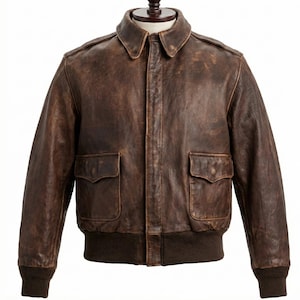 Può includere: Giacca bomber in pelle marrone dal design classico. La giacca presenta due tasche frontali, orlo e polsini a coste e un colletto. La giacca è esposta su un manichino.