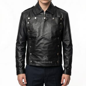 Puede incluir: Chaqueta de cuero negro con botones plateados y cierre de cremallera. La chaqueta tiene cuello y mangas largas con hebillas. El nombre de la marca "ROADHIDE" está impreso en la parte delantera.