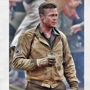 Op de afbeelding: Een bruine bomberjack met daaronder een donkergroen overhemd. De jas heeft een ritssluiting, twee voorzakken en een patch op de linkermouw. De persoon op de foto houdt een kleine beker vast.