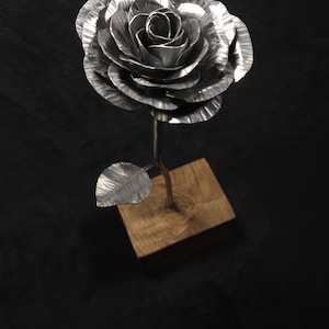 Peut inclure: Une sculpture de rose en métal avec des pétales argentés texturés et superposés, ainsi qu'une feuille unique sur une tige fine. La fleur est fixée sur un socle carré en bois brun clair, créant un contraste entre le métal et le bois.