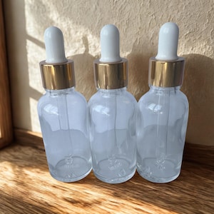 May include: Three clear glass dropper bottles with white droppers and gold-coloured collars. The bottles are empty and sit on a wooden surface. The background is a light-coloured wall and a wooden window frame.