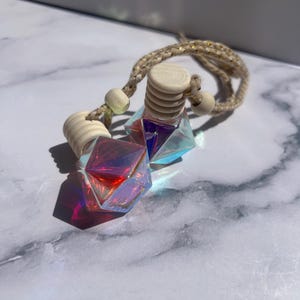 May include: Two iridescent car air fresheners with geometric crystal-like shapes. Each has a wooden cap and is suspended by a braided beige cord with wooden beads. The crystals reflect red, purple, and blue hues.