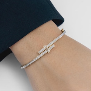 May include: A gold bangle bracelet featuring a row of small round pavé-set diamonds along the band. The center design consists of two overlapping horizontal bars, each accented with baguette-cut diamonds and a central rectangular stone.