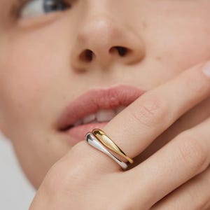 Könnte beinhalten: Nahaufnahme einer Hand mit zwei Ringen. Ein Ring ist goldfarben, der andere silberfarben. Die Ringe haben ein glattes, geschwungenes Design mit minimalistischer Ästhetik. Die Ringe befinden sich am Ringfinger.
