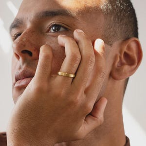 May include: A simple gold band ring with a smooth, matte finish. The ring features a flat, uniform profile and a consistent width, encircling the finger with a clean, metallic appearance that reflects light evenly across its surface.