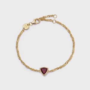 May include: A gold chain bracelet featuring a twisted link design and a single triangular red gemstone set in a gold bezel. The bracelet includes a lobster claw clasp and a small circular charm engraved with the letter D near the closure.