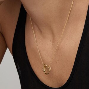 May include: A gold-toned necklace featuring a delicate chain and a pendant composed of three interlocking, smooth circular rings. The pendant hangs centrally, showcasing a polished metallic finish that reflects light across its curved surfaces.