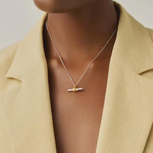 May include: A delicate silver chain necklace featuring a central pendant composed of a horizontal silver bar intersected by a small, polished gold-toned knot element. The necklace rests against the neck, showcasing a two-tone metallic design.