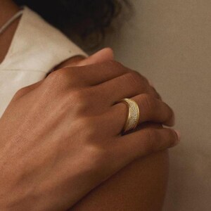 Könnte beinhalten: Ein breiter Goldring mit einer texturierten, schimmernden Oberfläche in der Mitte. Der Ring hat ein gewölbtes, gebogenes Profil und wird am Ringfinger getragen, wobei er ein poliertes Metallfinish aufweist, das das Licht auf der Oberfläche reflektiert.