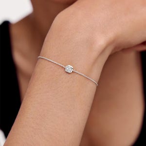 May include: A delicate silver-toned chain bracelet featuring a single, small, round clear gemstone set within a square-shaped metal bezel. The bracelet is thin and minimalist, with the stone positioned centrally along the fine link chain.
