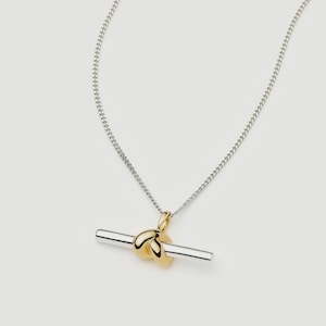 May include: A silver chain necklace featuring a toggle-style pendant. The pendant consists of a polished gold-tone knot element centred on a smooth, horizontal silver-tone bar. The design combines metallic finishes in a simple, clean aesthetic.