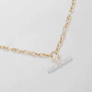 May include: A gold chain necklace featuring a toggle clasp closure. The toggle bar is cylindrical and encrusted with small, clear, sparkling stones. The chain consists of interconnected oval links with a textured, polished gold finish throughout.