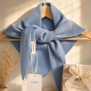 May include: A light blue triangular knit scarf draped over a wooden hanger. A white tag attached to the scarf displays the text 100%WOOL and ELVA ROSE. Beside it sits a gift box wrapped in tan paper with a cream ribbon and a card reading A little gift for you.