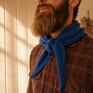 May include: A bright blue knit scarf tied in a simple knot around the neck, worn over a brown and black plaid button-down shirt with two chest pockets and dark buttons. The scarf features a textured, soft-looking knit pattern.