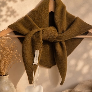 May include: A triangular olive green knit scarf made of 100% wool, draped over a wooden hanger. A white fabric tag attached to the scarf reads 100% WOOL, and a separate white rectangular paper tag features the text ELVA ROSE in black lettering.