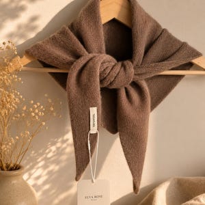 May include: A brown triangular knit scarf made of 100% wool, draped and knotted on a wooden hanger. A white fabric tag attached to the scarf reads 100% WOOL, and a separate white rectangular brand tag displays the text ELVA ROSE Boutique.