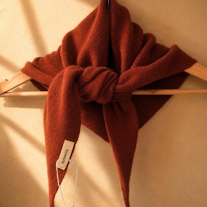 May include: A rust-coloured triangular scarf made of knit fabric, tied in a central knot and draped over a wooden hanger. A small rectangular fabric tag attached to one end of the scarf features the text 100% WOOL printed in black ink.