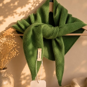 May include: A triangular green knit scarf made of 100% wool, draped over a wooden hanger. A rectangular fabric tag attached to the scarf reads 100% WOOL, and a white paper tag hanging from it displays the text ELVA ROSE Boutique.
