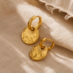 May include: A pair of gold-toned hoop earrings featuring circular pendants. Each pendant is embossed with a crescent moon, radiating sun rays, and small star motifs. The earrings have a polished finish and a hinged closure mechanism for secure wear.