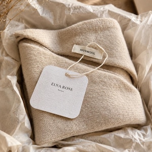 May include: A folded beige wool garment inside a box lined with white tissue paper. A white rectangular tag attached with a string displays the text ELVA ROSE Boutique, and a smaller fabric label attached to the garment reads 100% WOOL.