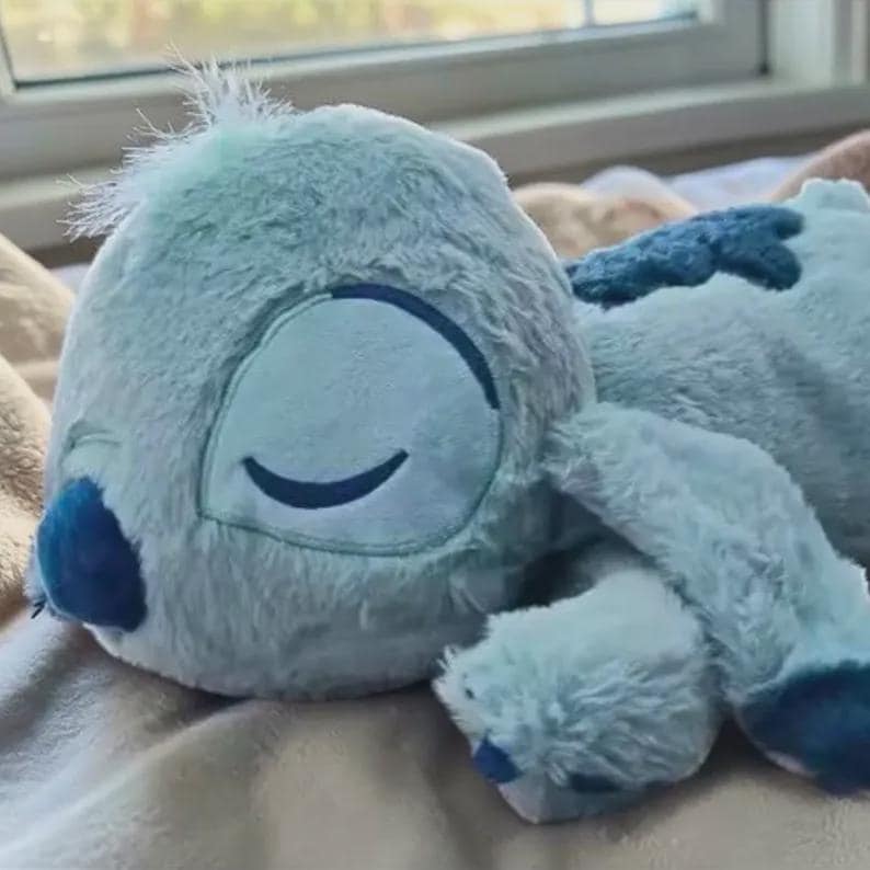 Puede incluir: Un peluche azul claro con forma de personaje de dibujos animados, tumbado sobre una superficie suave. El juguete tiene ojos grandes y cerrados, una nariz azul oscuro y detalles azul oscuro en las orejas y la espalda. El peluche es suave y esponjoso.