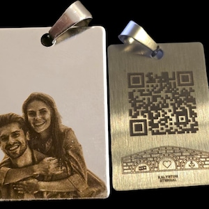 Può includere: Due pendenti rettangolari in metallo con anelli color argento. Uno presenta un'immagine incisa di una coppia sorridente. L'altro pendente ha un codice QR e un disegno di ponte inciso con il testo "KALYTON HTIRIAL".