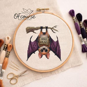 May include: A cross-stitch embroidery hoop featuring a bat design hanging upside down from a branch. The bat has purple wings and the text "FREE HUGS". Embroidery floss in various colors and a pair of small gold scissors are also visible.